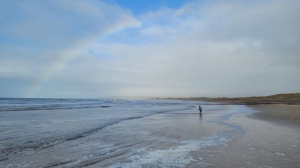 The beach of Westport on the Mull of Kintyre. A small child in the distance splashes through the shallow waves. A half rainbow covers the sky above the sand dunes.